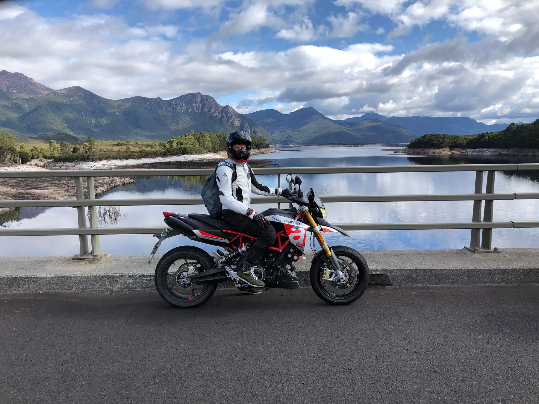 A helmeted rider on a motorcycle pauses by a railing above the water, seen at a distance.