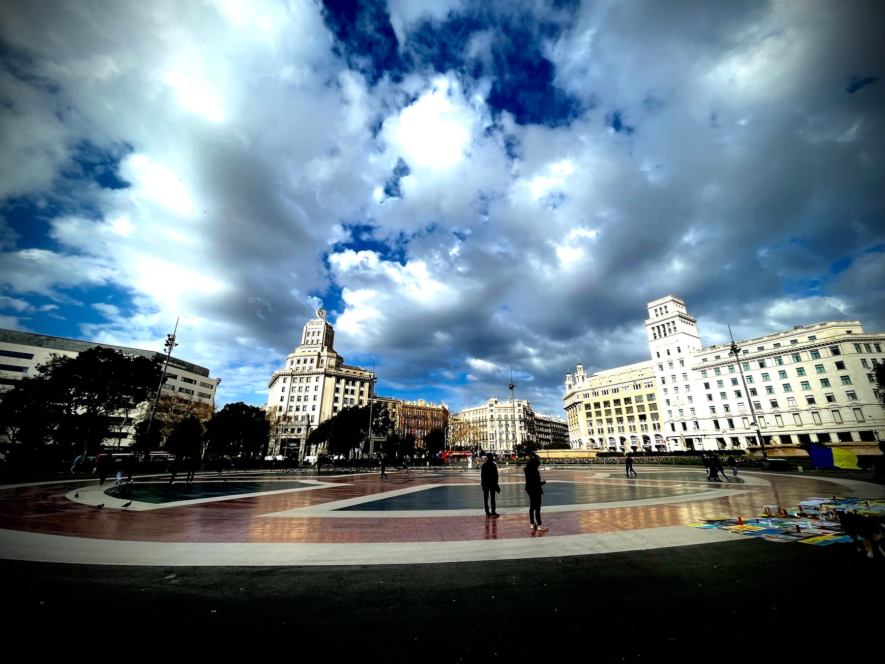 A wide city plaza beneath a vast sky of broken cloud, a low fountain in the foreground and grand buildings beyond.