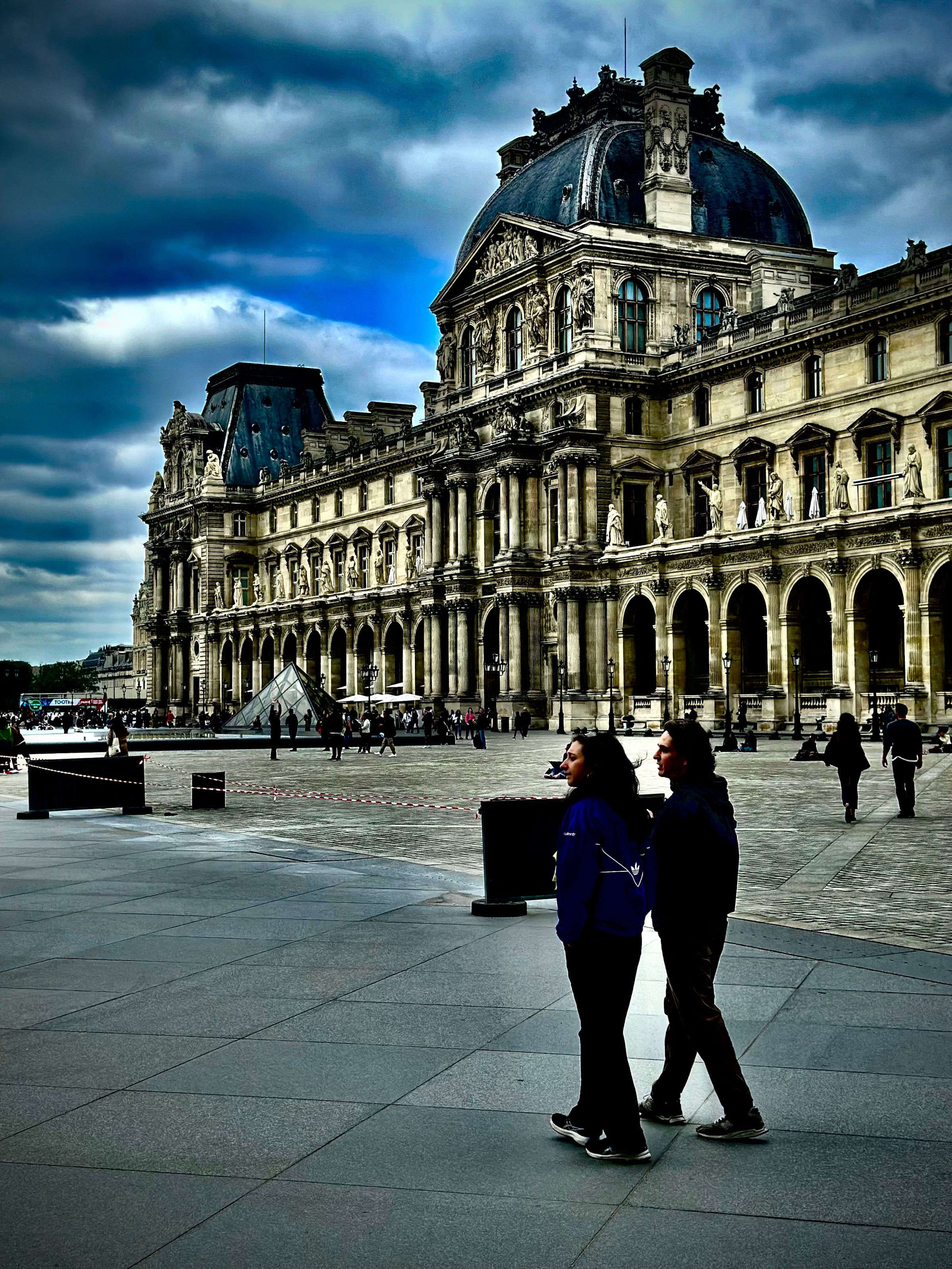 A grand palace courtyard under a heavy sky, a couple walking across the open paving in the distance.