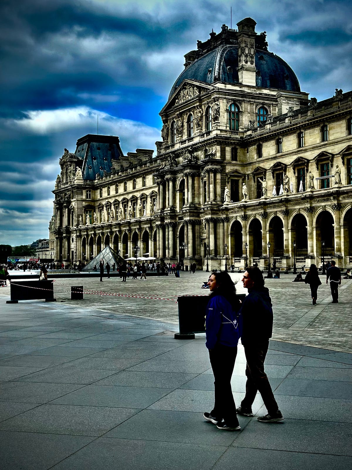 A grand palace courtyard under a heavy sky, a couple walking across the open paving in the distance.