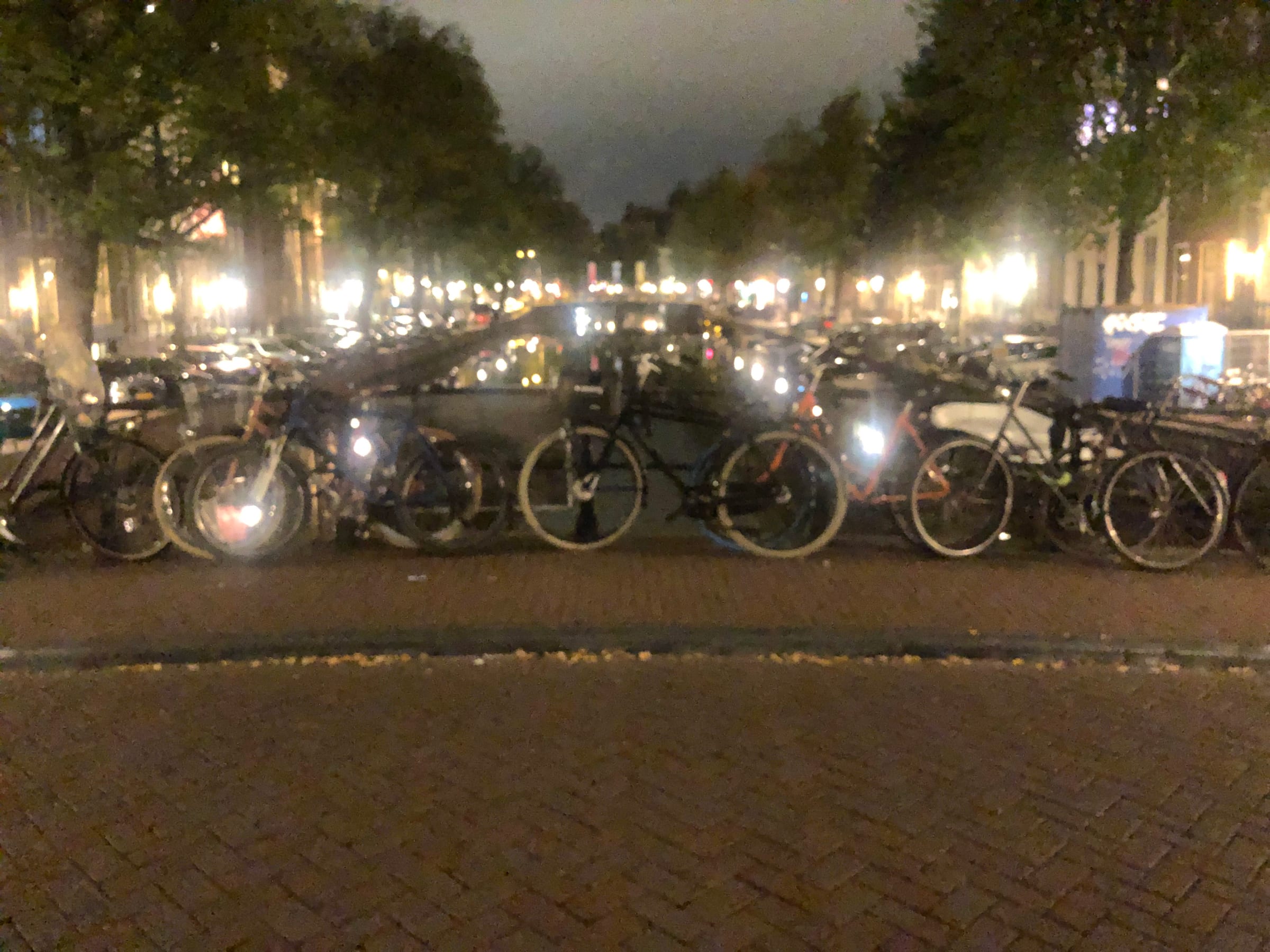 Parked bicycles crowded along a canal railing at night, their forms blurred under warm street light.