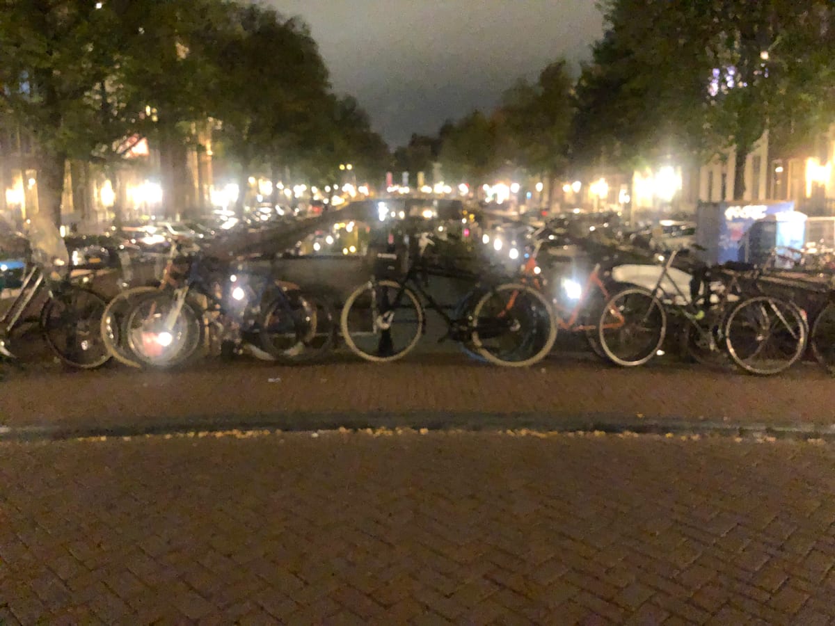 Parked bicycles crowded along a canal railing at night, their forms blurred under warm street light.