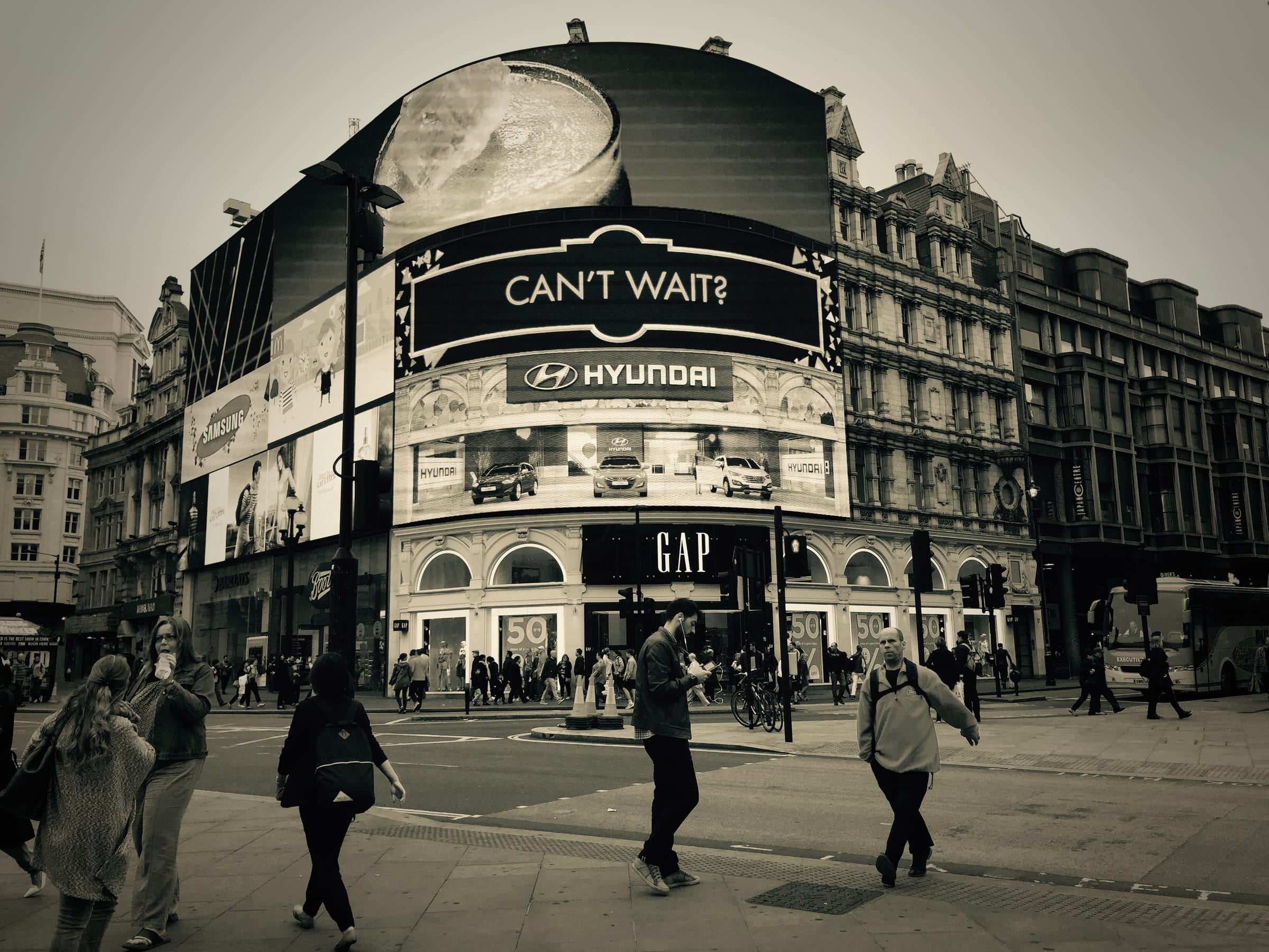 A busy city junction ringed by bright advertising screens, a crowd crossing beneath the hoardings.