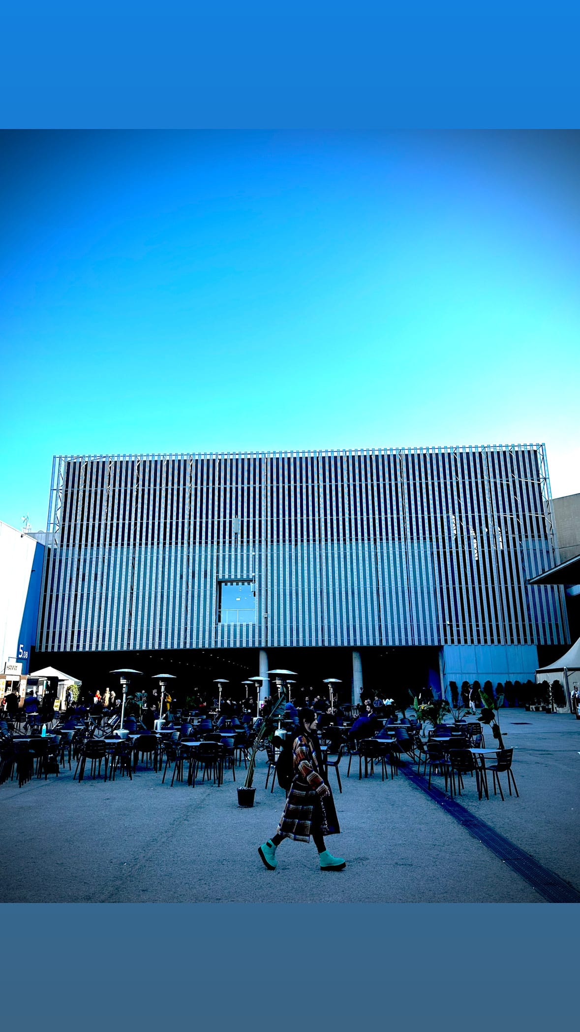 A pale modernist plaza under clear blue sky, one distant figure walking across the open space.