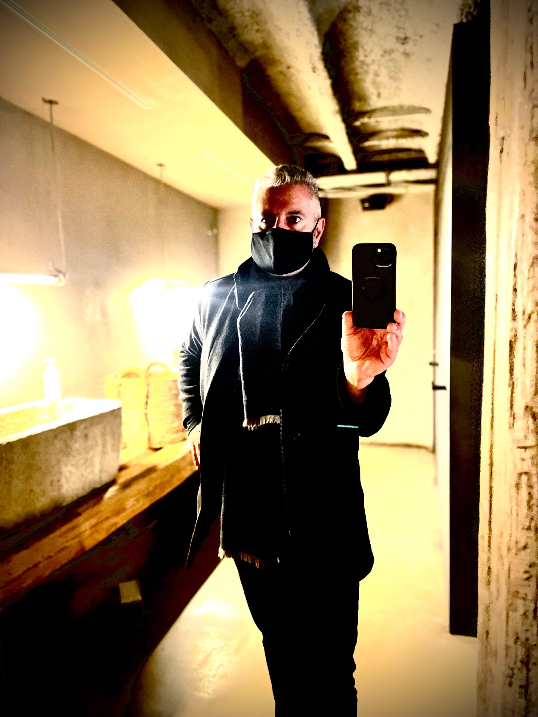 A masked figure photographs themselves in a mirror along a warm-lit corridor.