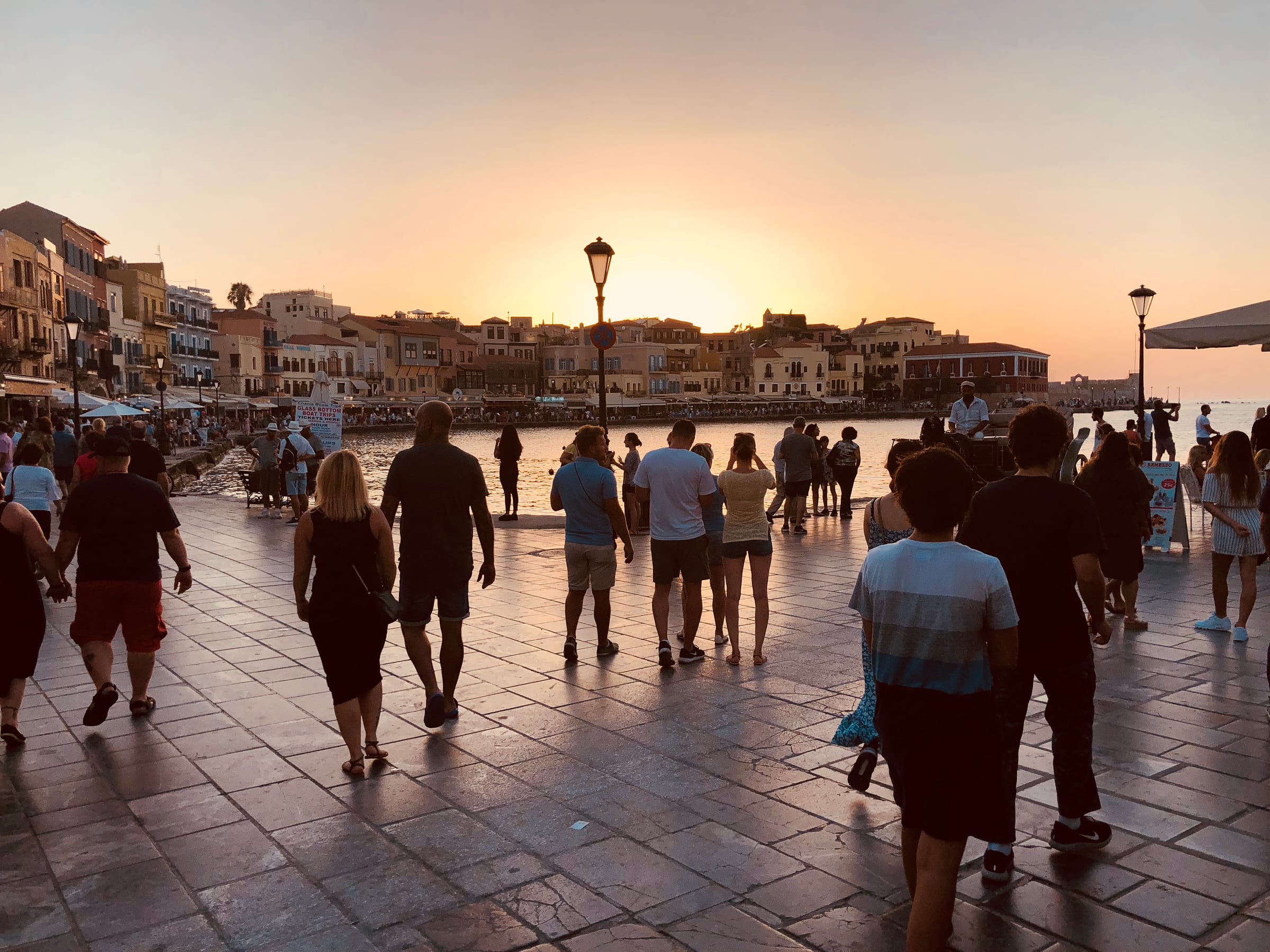 A harbour promenade at sunset, a crowd of people walking away along the waterfront, the sky burning orange.