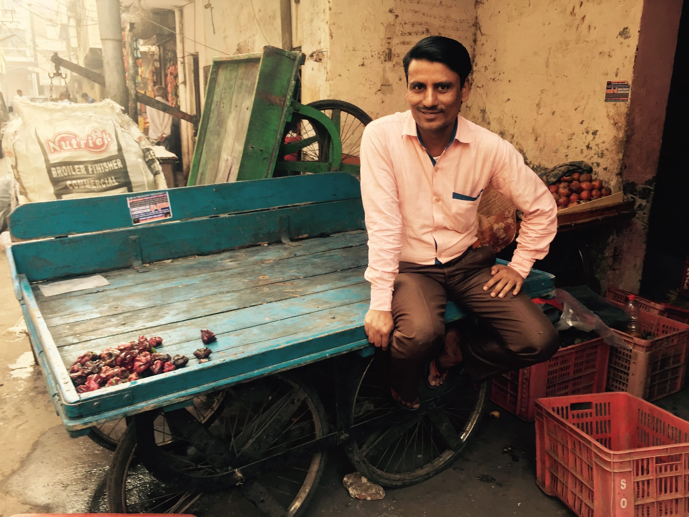 A young man stands beside a blue wooden cart in a bright street.