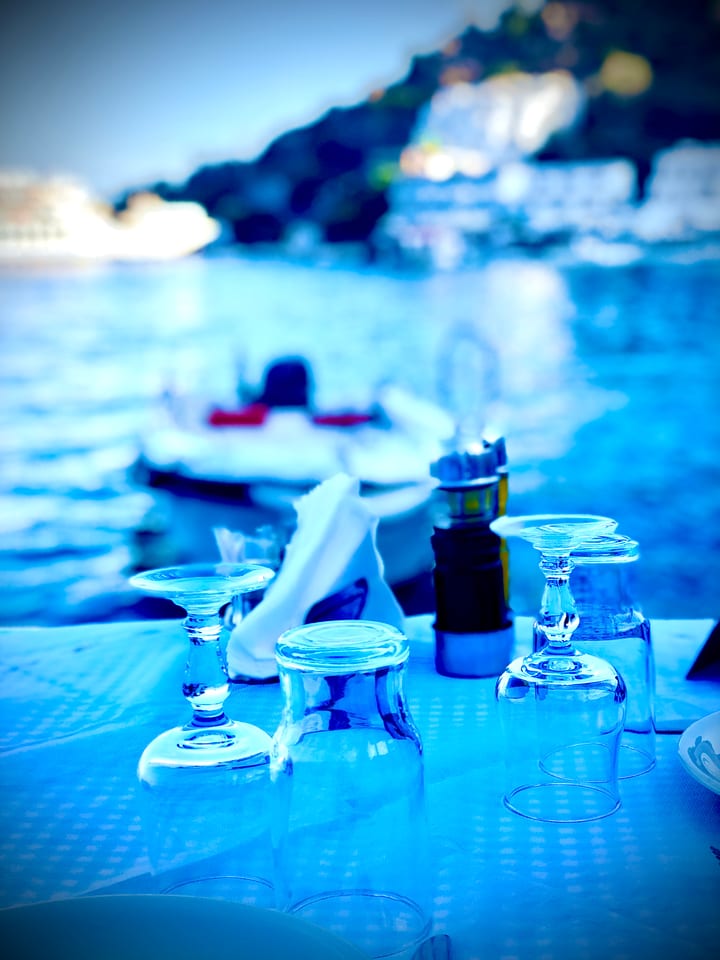 A taverna table set with glasses by the sea at blue hour, the water softly out of focus behind.