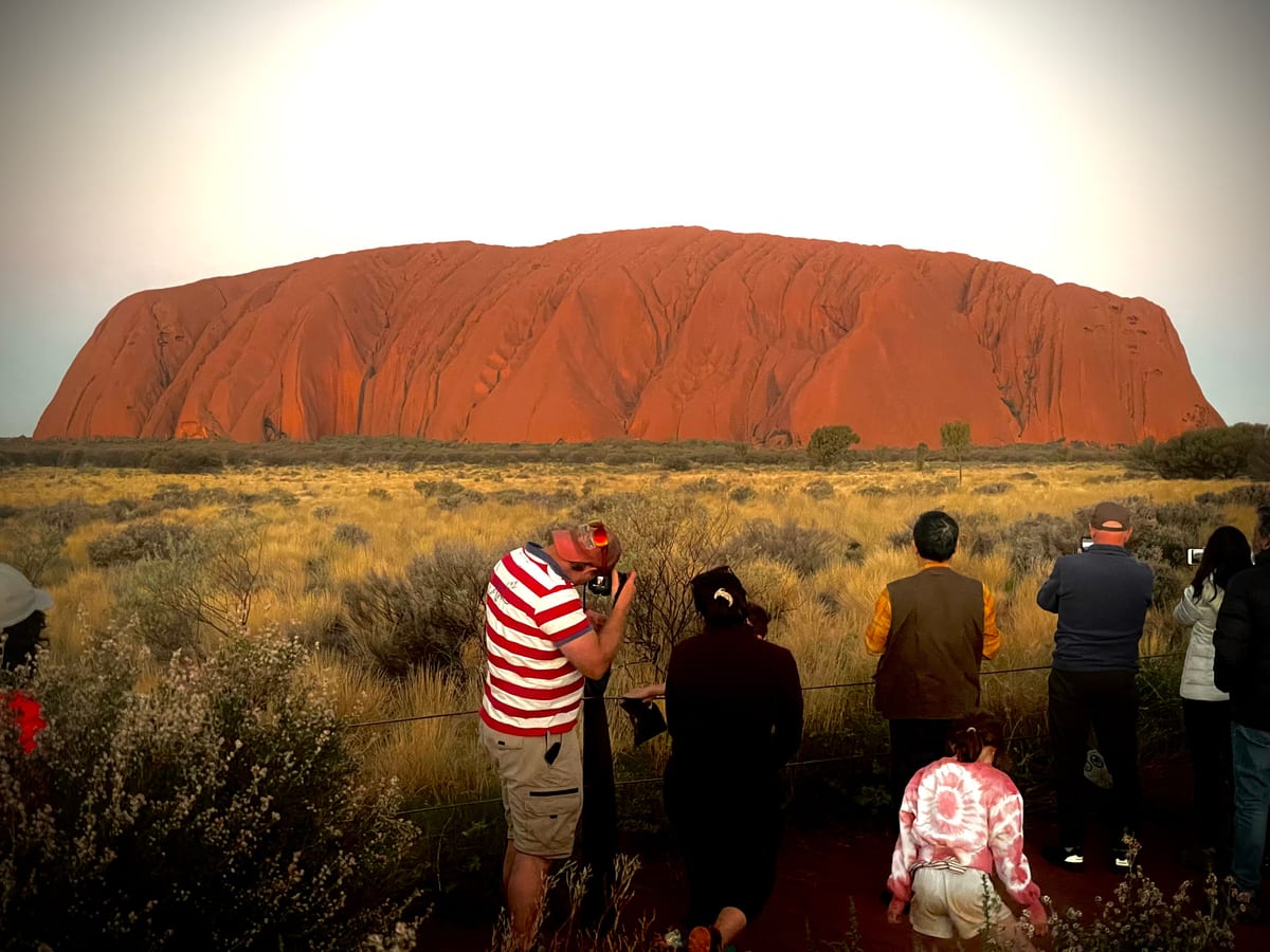 A group of onlookers, seen from behind, watch a great red monolith glow at sunset across the scrub.
