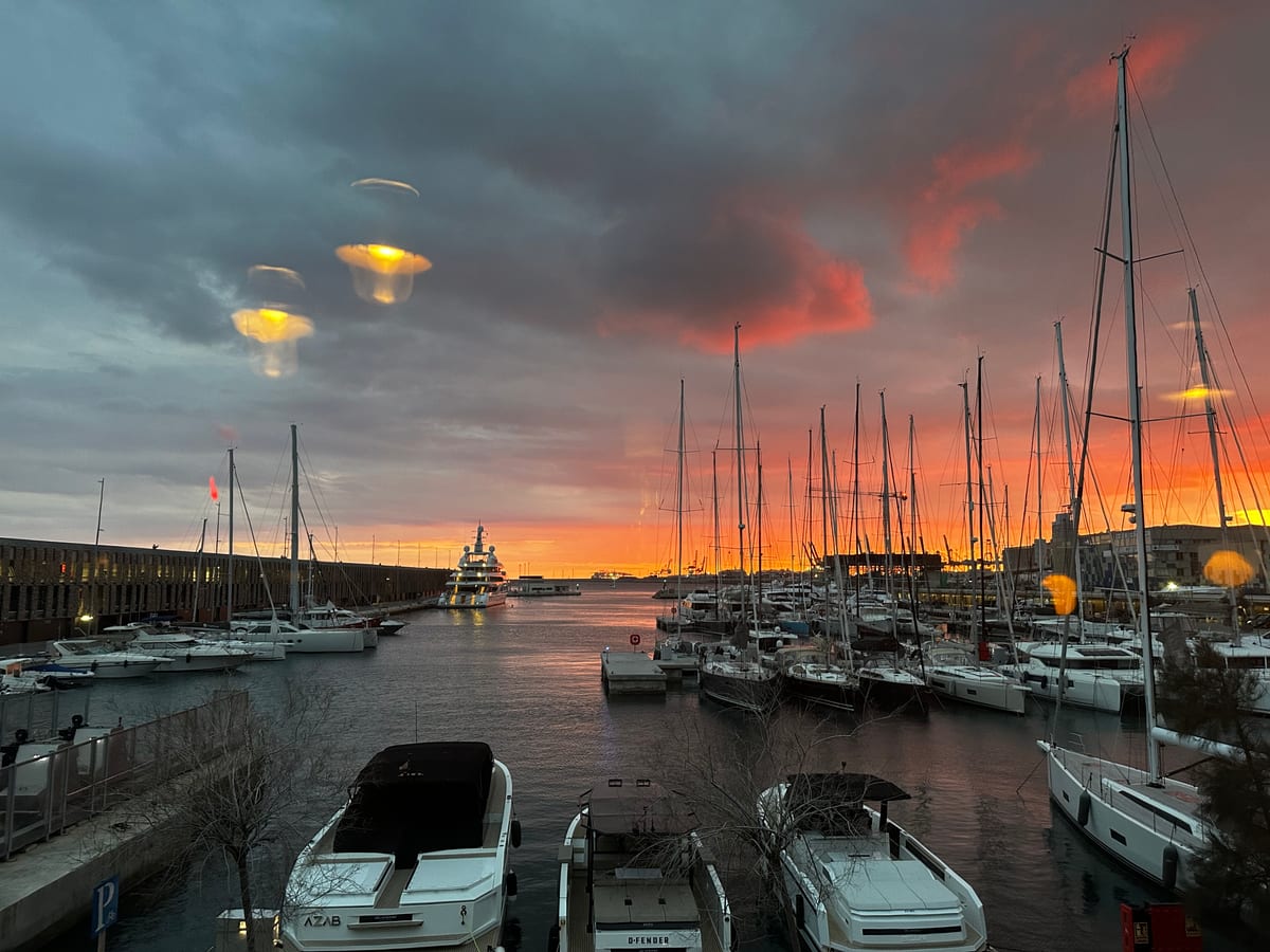 A marina at sunset, yacht masts silhouetted against an orange and grey sky, parked cars along the quay.