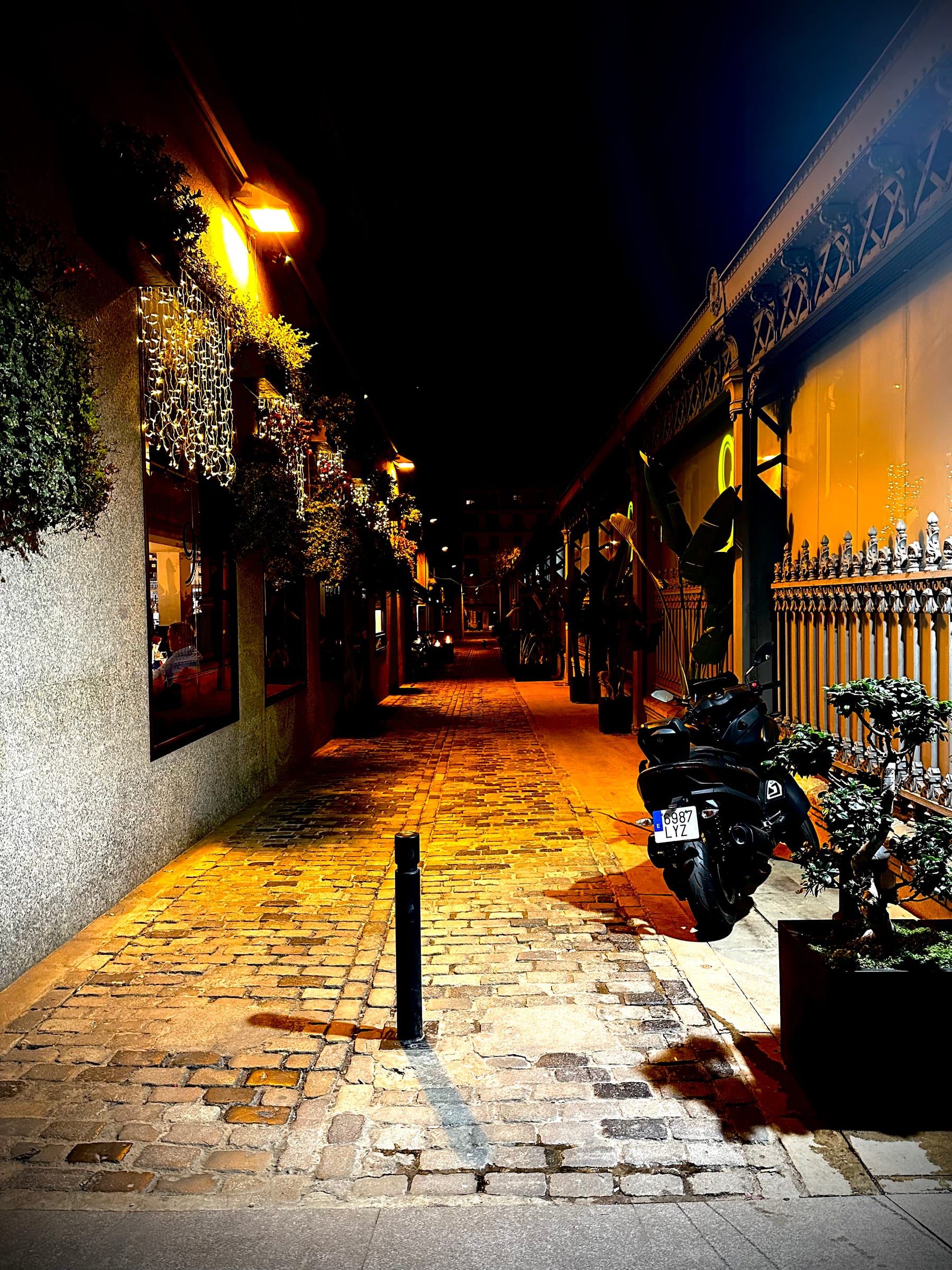 A narrow alley at night lit by warm street lamps, wet cobblestones reflecting the glow.