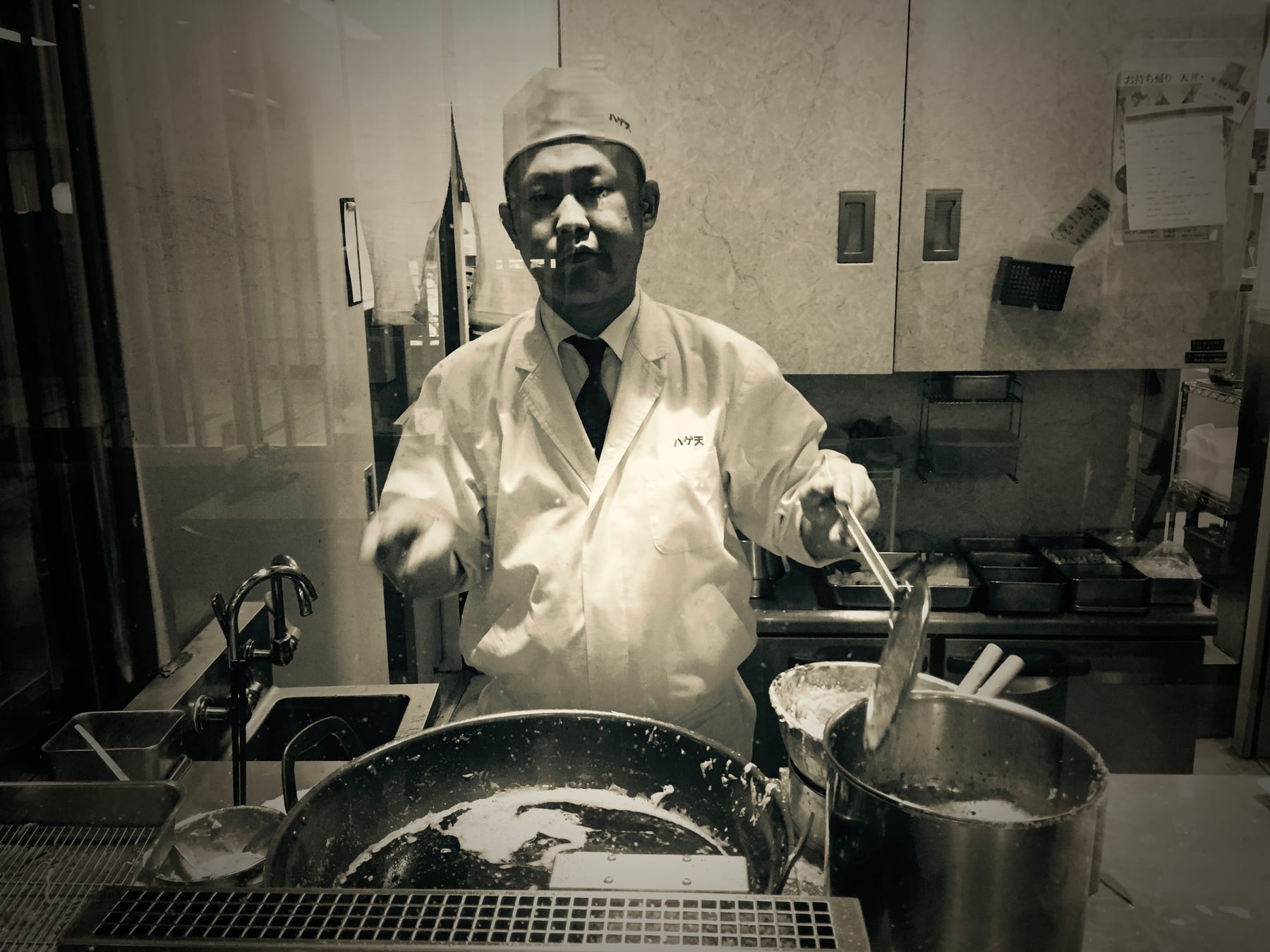 A cook in whites works over a steaming pot in a kitchen, in black and white.