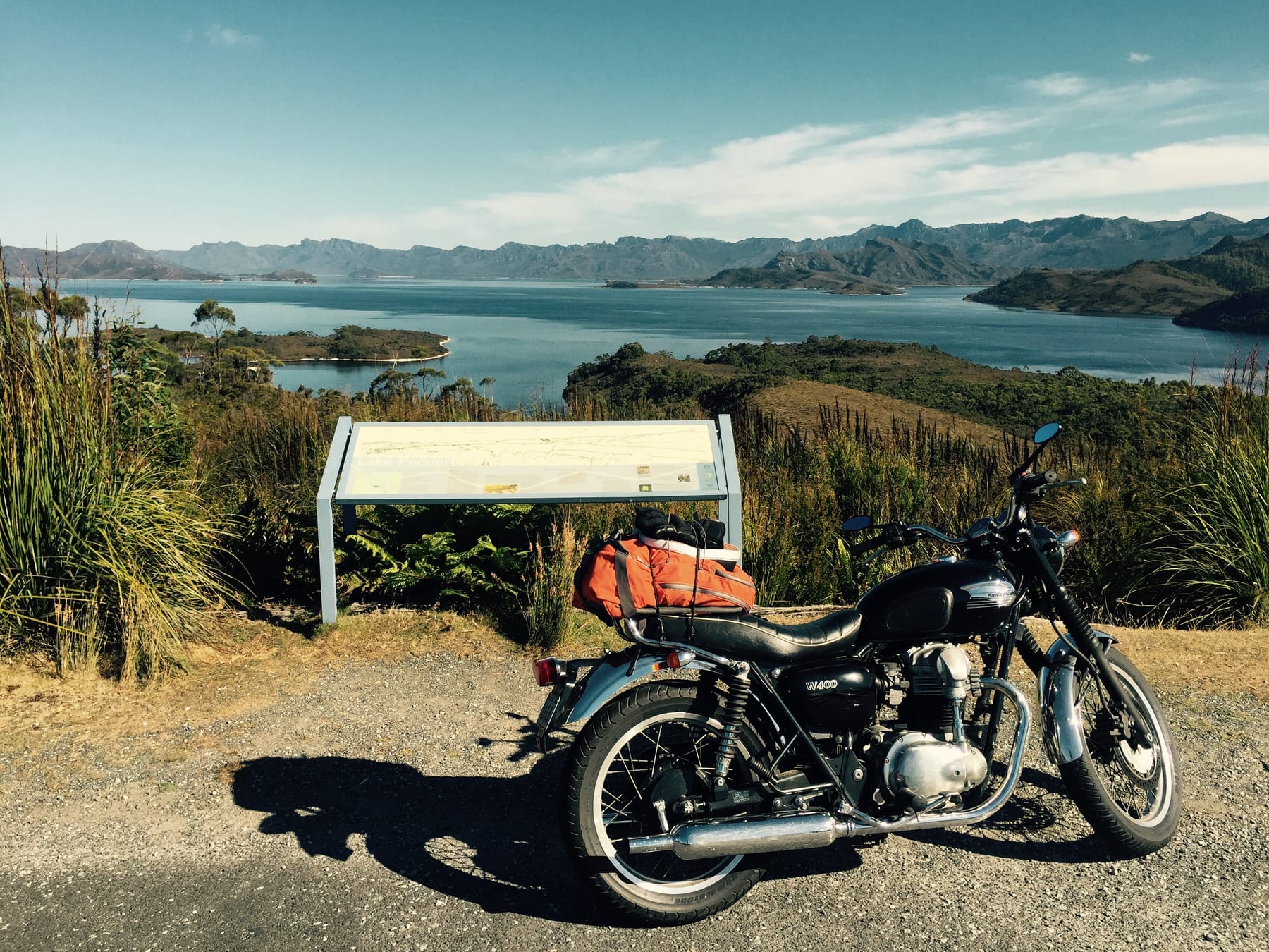 A classic motorcycle parked at a lookout above a calm bay ringed by hills.