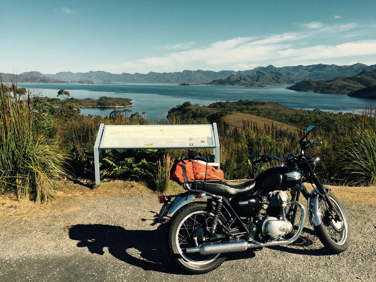 A classic motorcycle parked at a lookout above a calm bay ringed by hills.