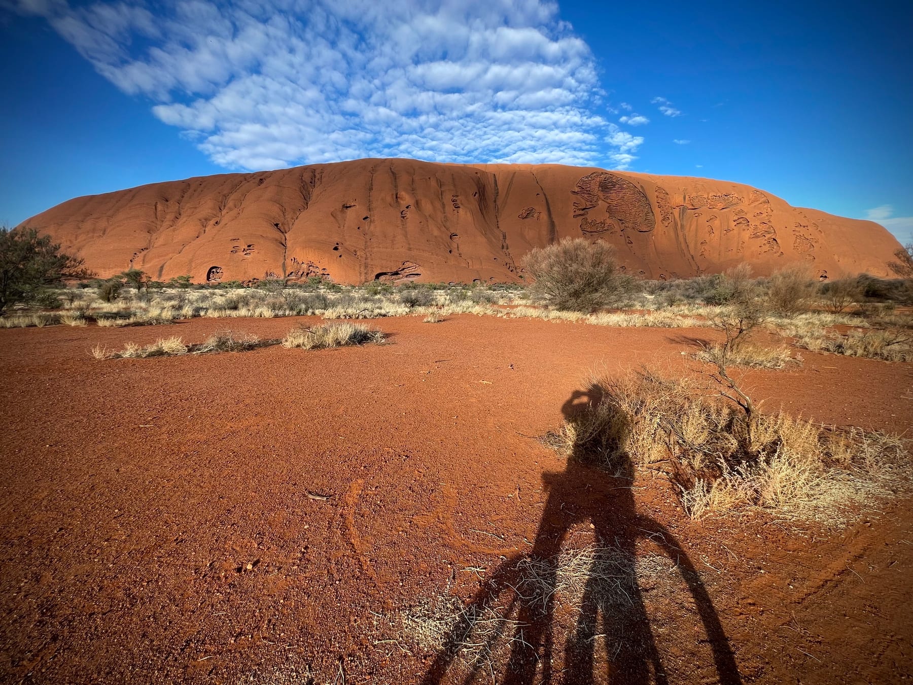 Red sand and scrub before a great rock face, the photographer's own long shadow falling into the frame.