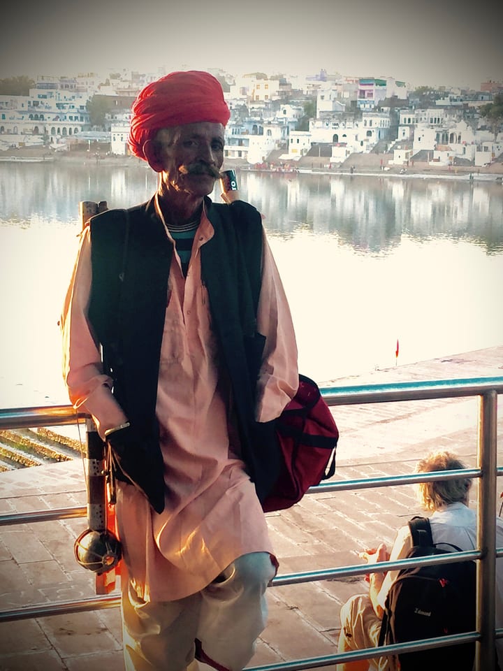 An older man in a red turban, seen in profile against a pale riverside.