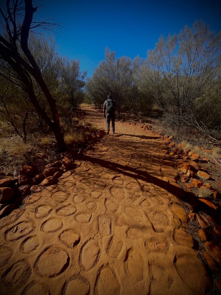 A red earthen walking path winding through desert scrub, a tiny distant figure on the trail.