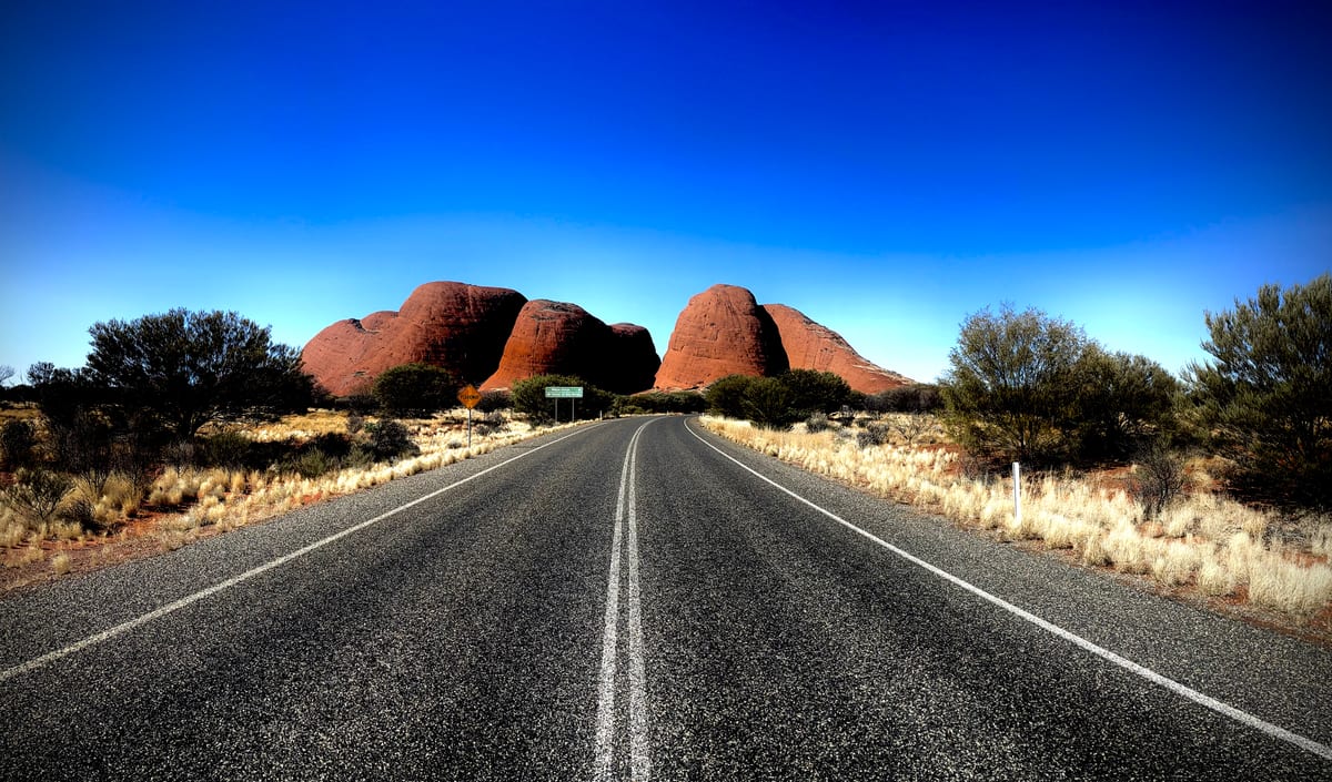 A long straight desert road running toward distant red rock domes under a deep blue sky.