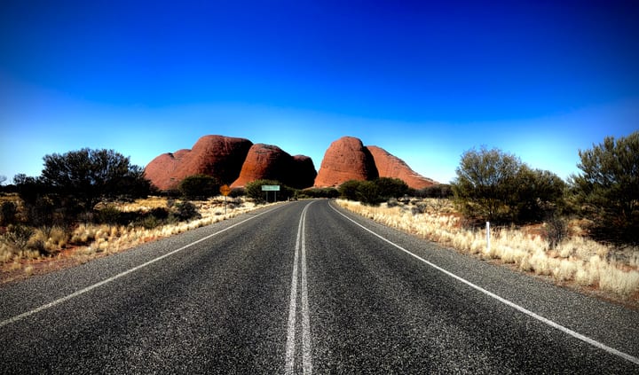 A long straight desert road running toward distant red rock domes under a deep blue sky.