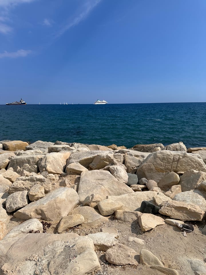A rocky breakwater meeting deep blue sea, faint boats on the far horizon.