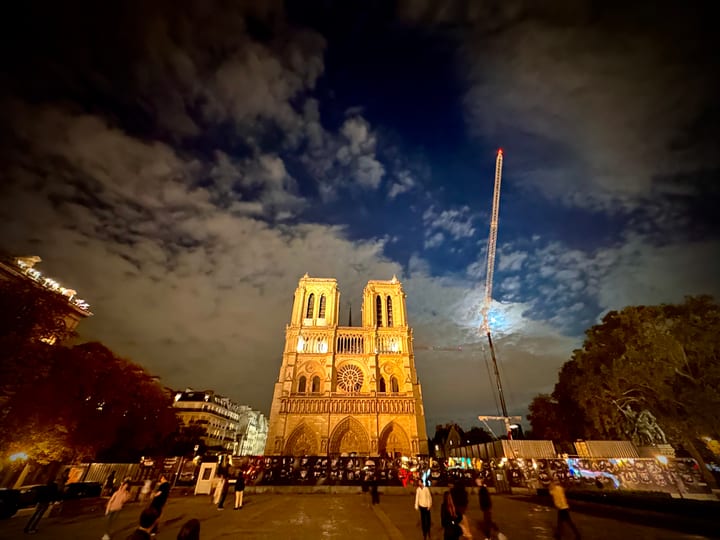 A great cathedral floodlit at dusk beneath a dramatic sky, a construction crane rising beside its towers.