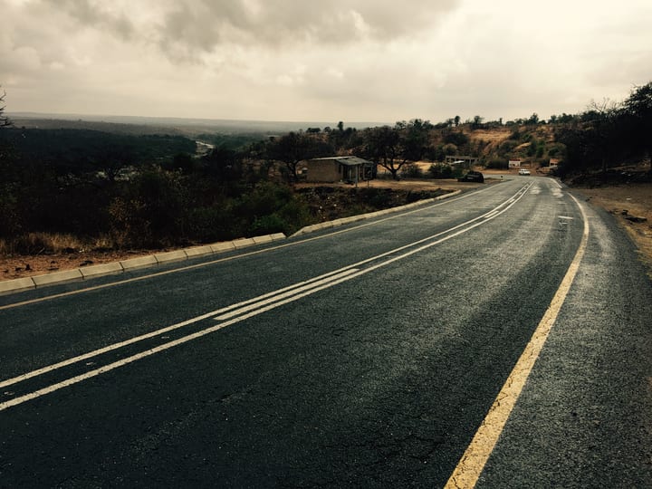 An empty tar road curving over a rise into open grassland under a grey, weather-heavy sky.