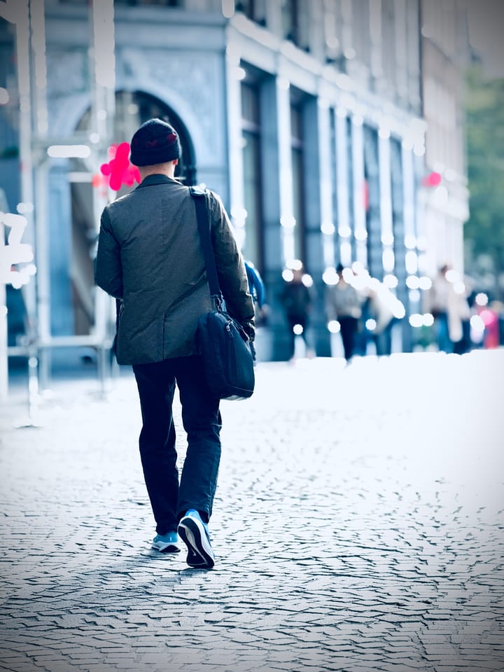 A lone figure with a backpack walks away down a pale stone street, seen from behind.