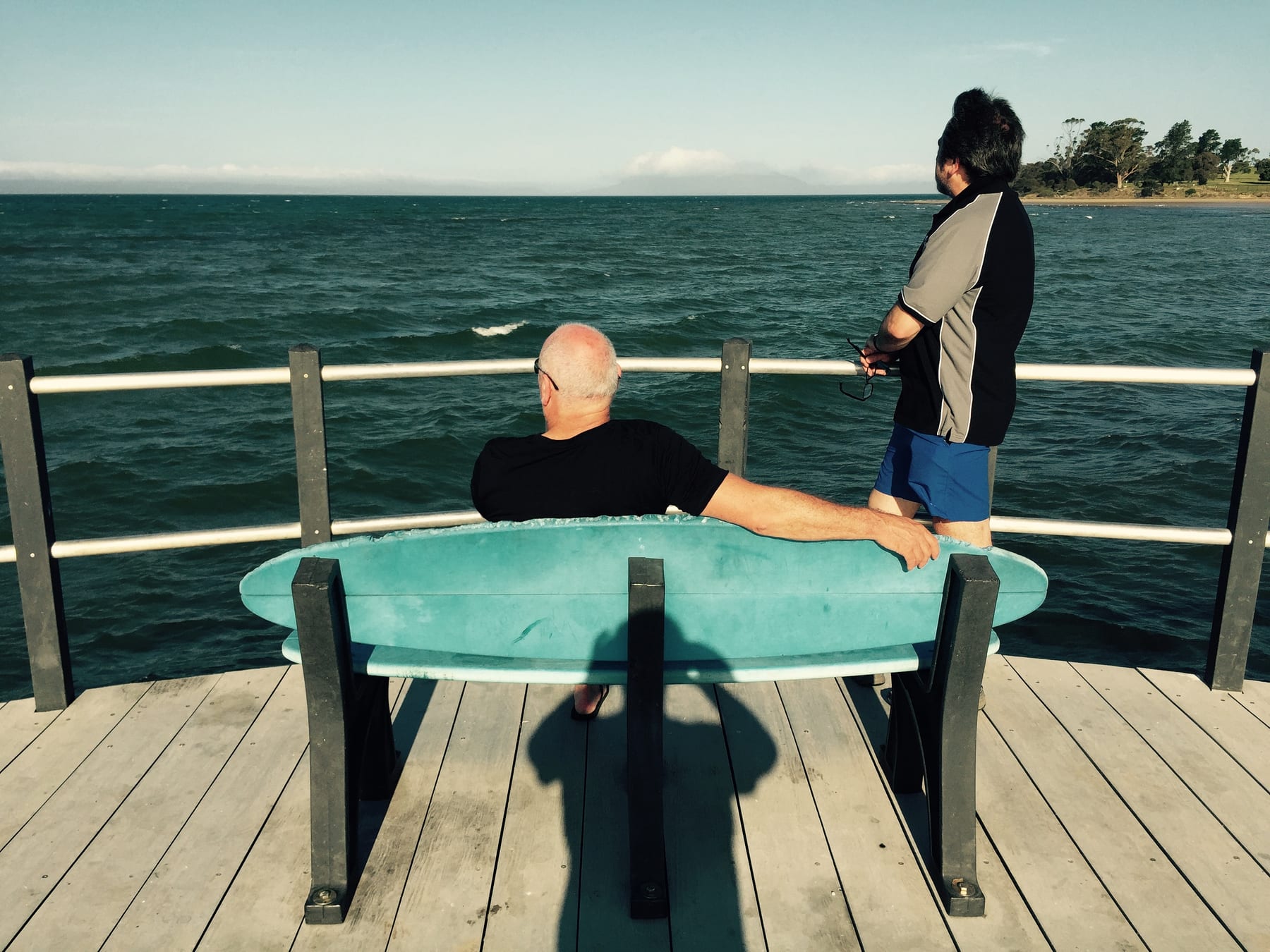 Two people rest on a bench on a timber deck above calm water.