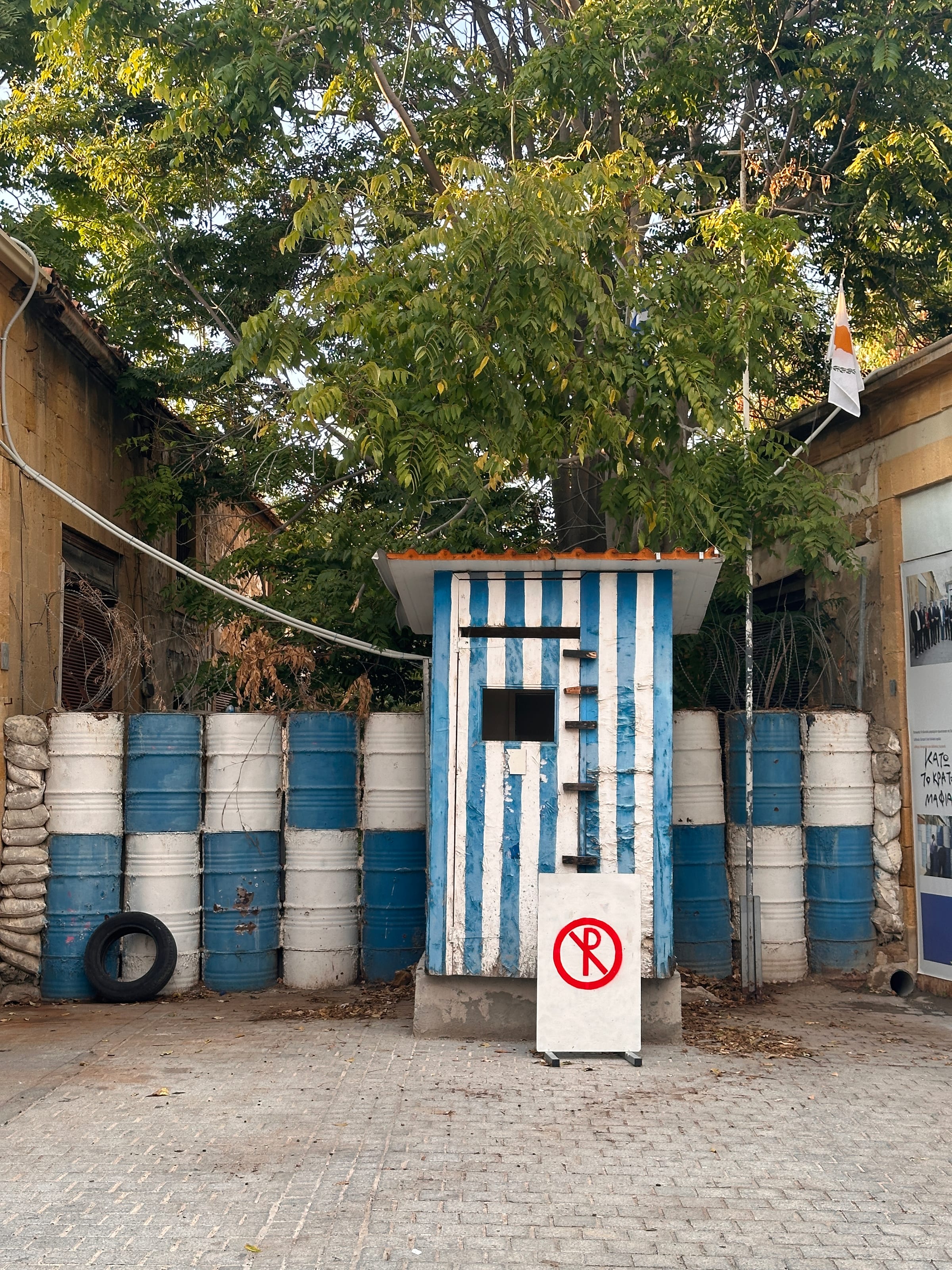 Blue and white painted oil drums standing under a tree beside a rough wall, a discarded tyre nearby.