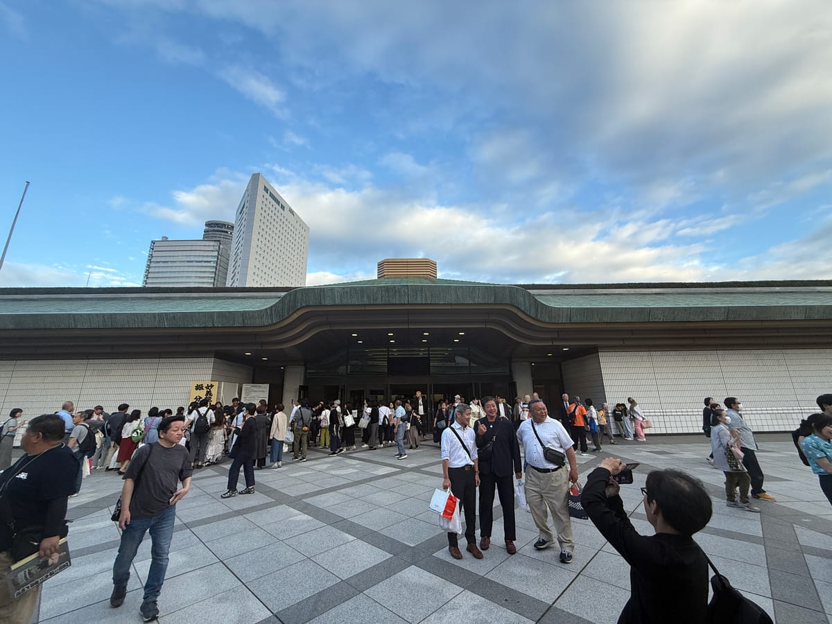 A broad station forecourt under a tall sky, a distant crowd gathered near the entrance.