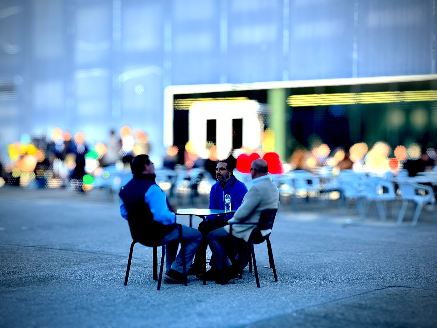 Two men sit talking at an outdoor cafe table, the background softly blurred.