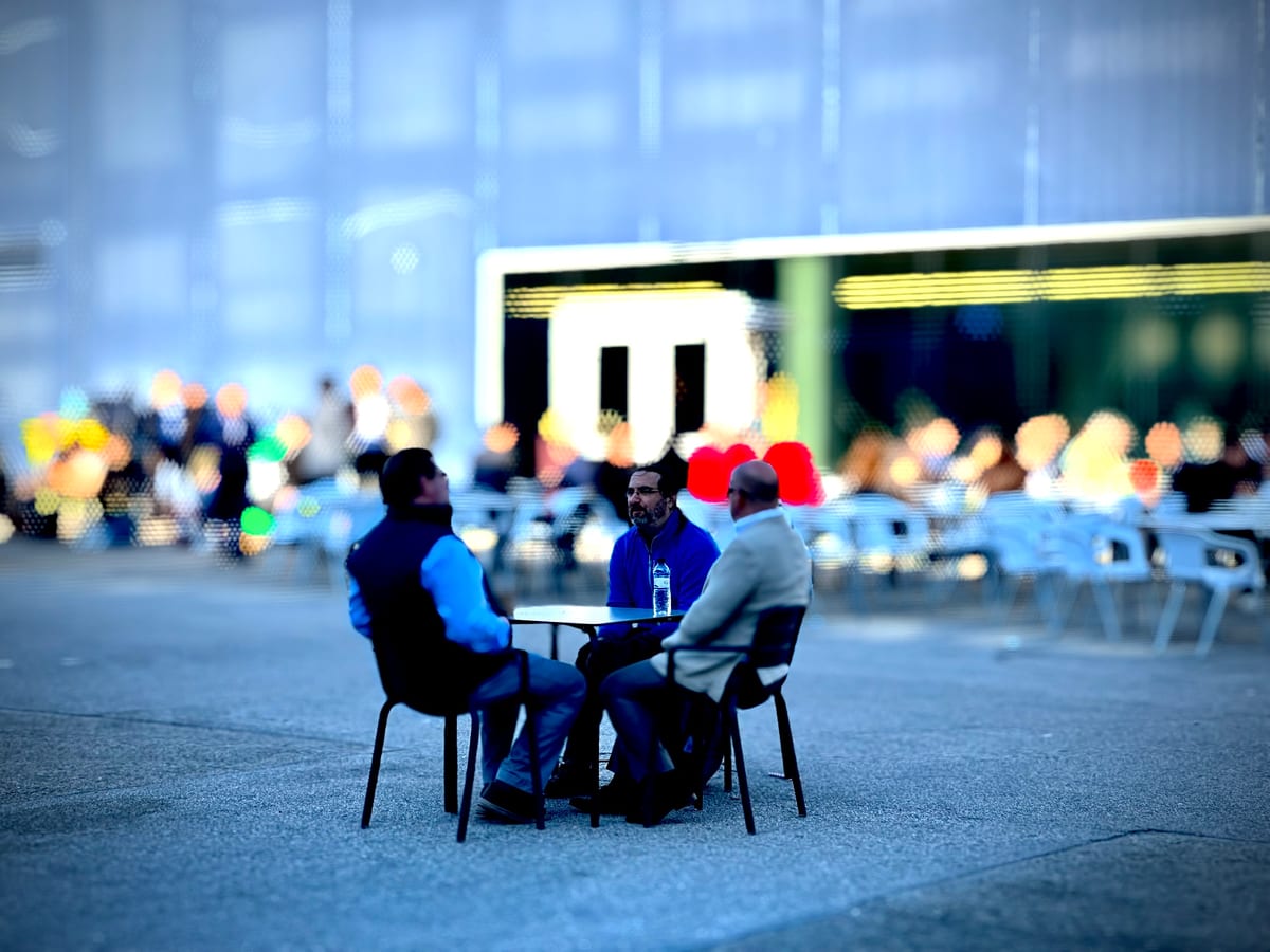 Two men sit talking at an outdoor cafe table, the background softly blurred.