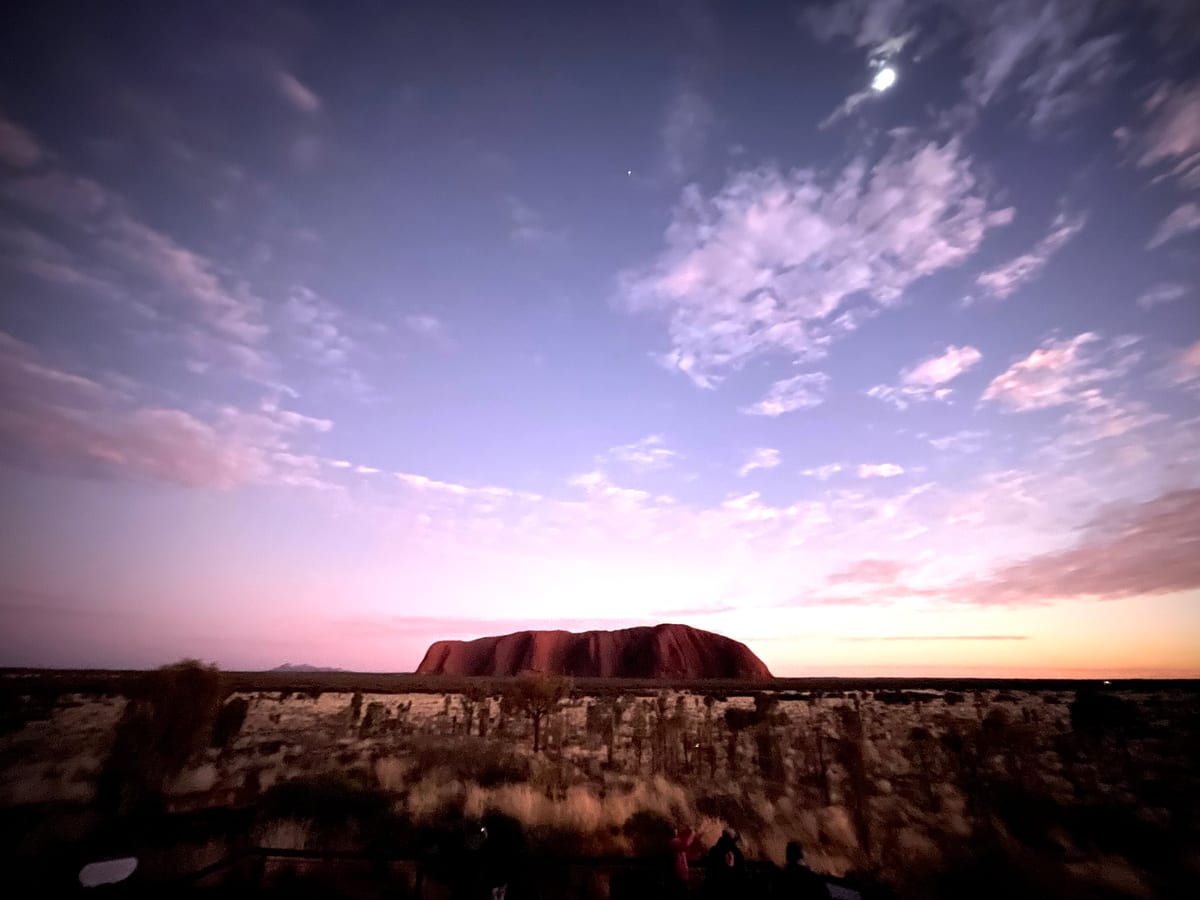 A red monolith low on the horizon at dusk beneath a wide lilac sky streaked with cloud.