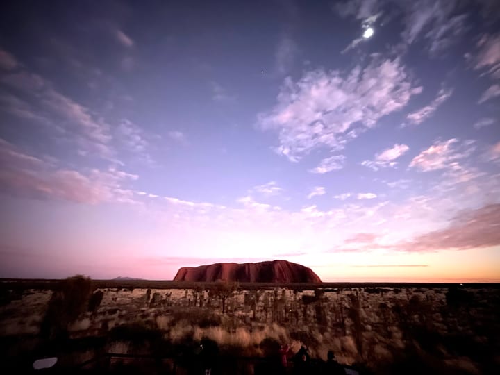 A red monolith low on the horizon at dusk beneath a wide lilac sky streaked with cloud.