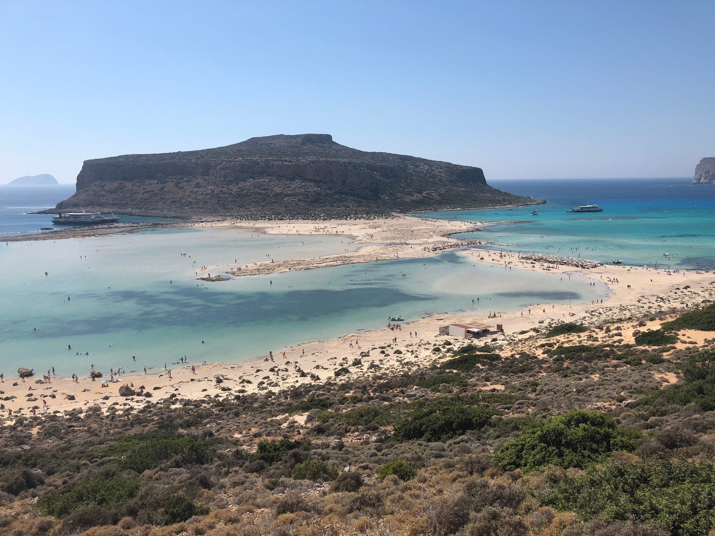 A shallow turquoise bay meeting a sweep of white sand and dark hillside, the open sea beyond.
