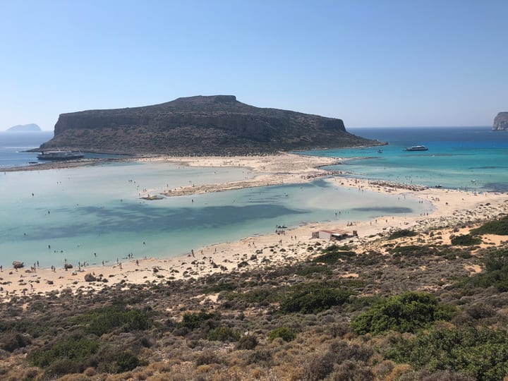 A shallow turquoise bay meeting a sweep of white sand and dark hillside, the open sea beyond.