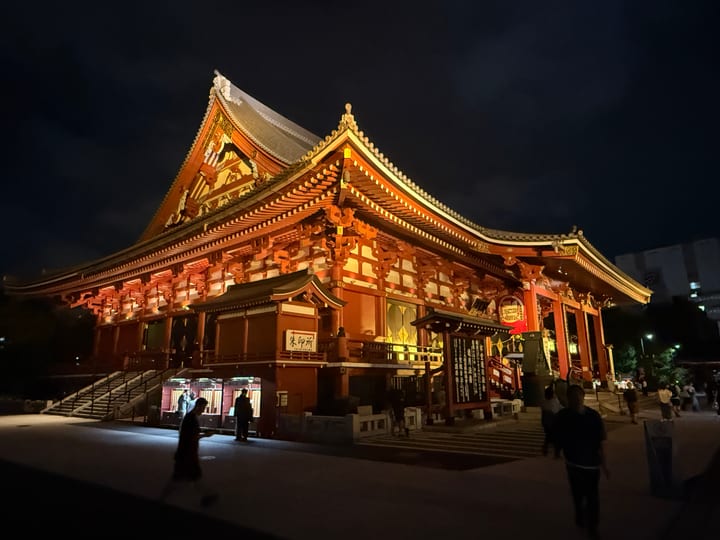 A red-and-gold temple hall lit at night, its lanterns glowing, a few small distant figures on the steps.