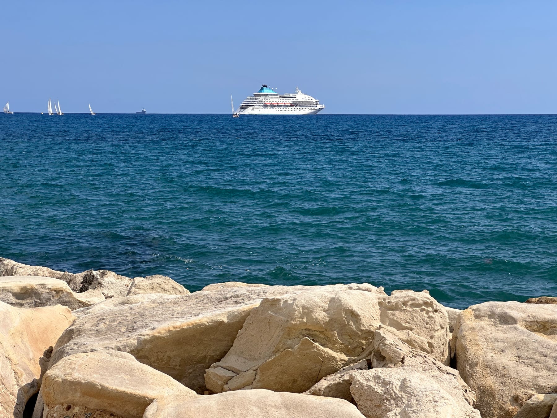 Open sea under a clear sky with a single white boat on the horizon, sun-bleached rocks in the foreground.