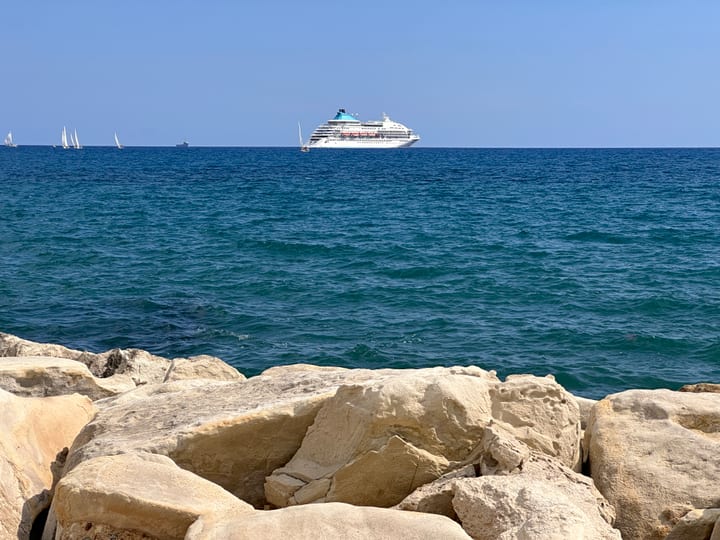 Open sea under a clear sky with a single white boat on the horizon, sun-bleached rocks in the foreground.