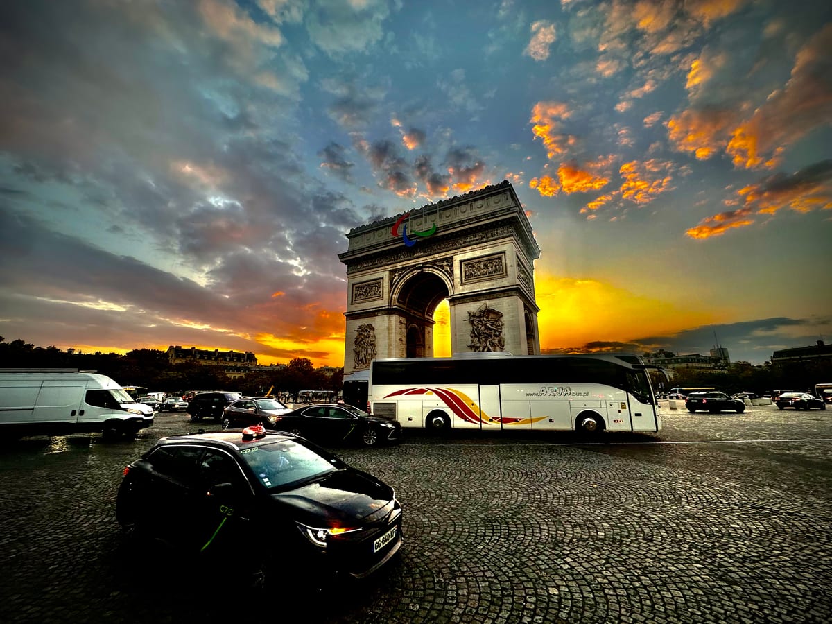 A triumphal arch at sunset above a busy roundabout, traffic and a coach in the foreground beneath blazing cloud.