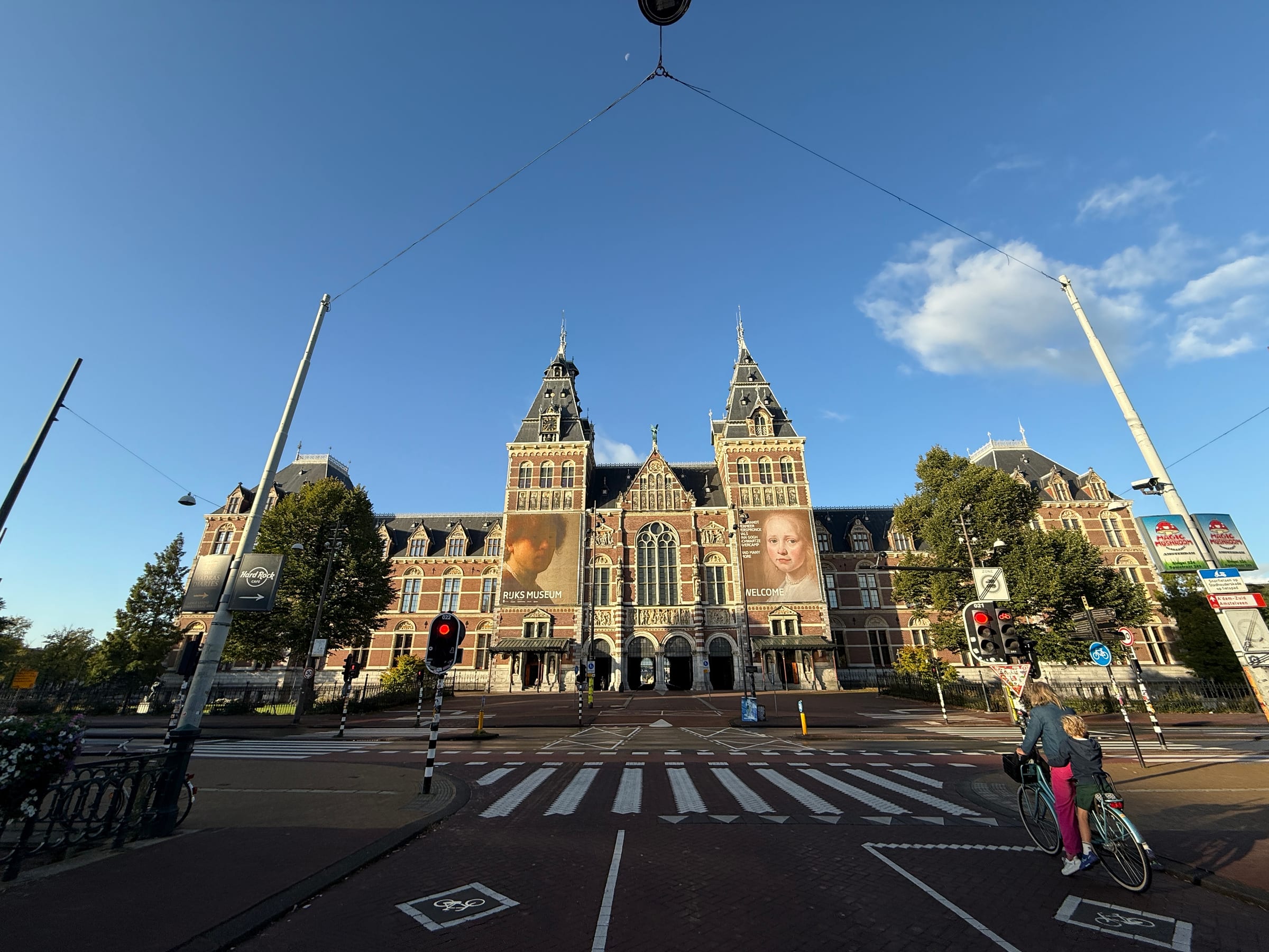 The Rijksmuseum seen across an empty forecourt under a bright blue sky, a single distant cyclist crossing.