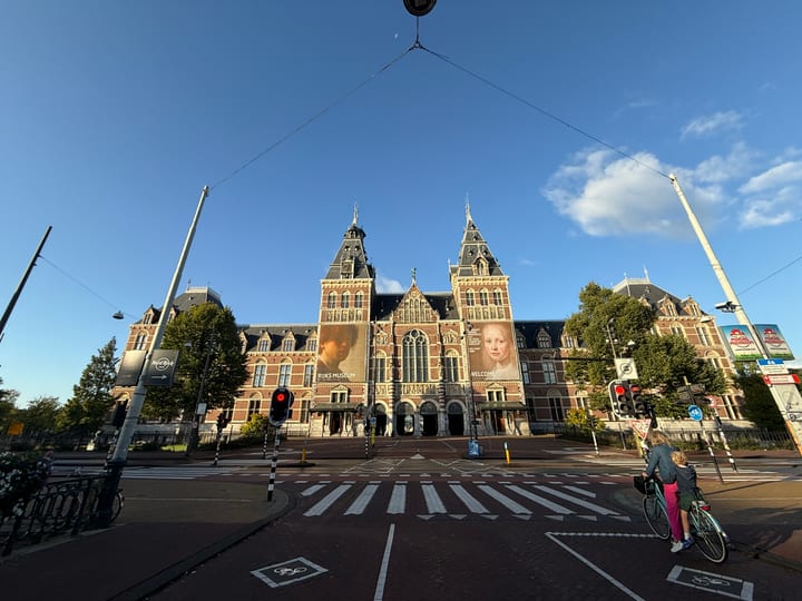 The Rijksmuseum seen across an empty forecourt under a bright blue sky, a single distant cyclist crossing.
