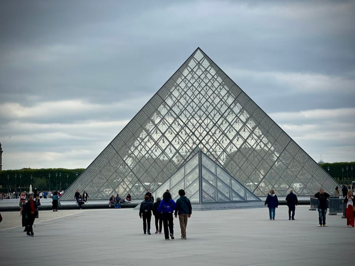 A glass pyramid in a museum courtyard beneath brooding cloud, scattered visitors small around its base.