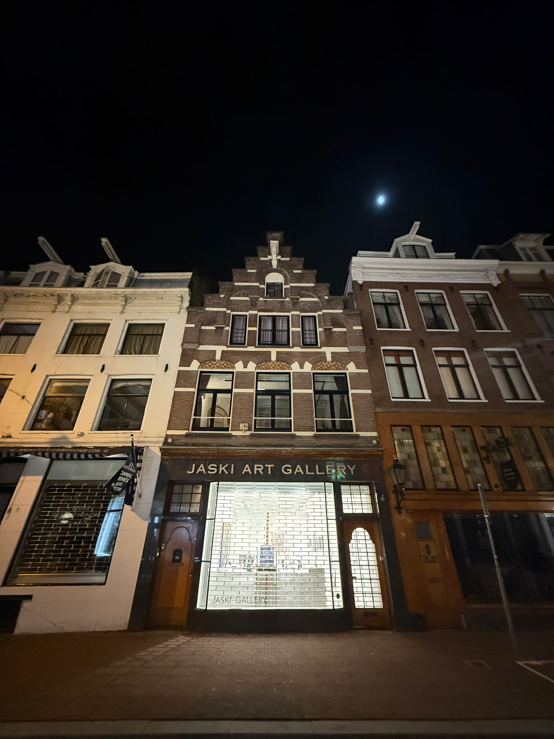 Tall narrow Amsterdam canal houses at night beneath a small bright moon, a lit gallery window at street level.