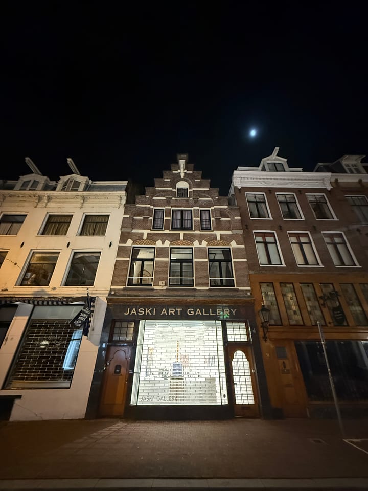 Tall narrow Amsterdam canal houses at night beneath a small bright moon, a lit gallery window at street level.