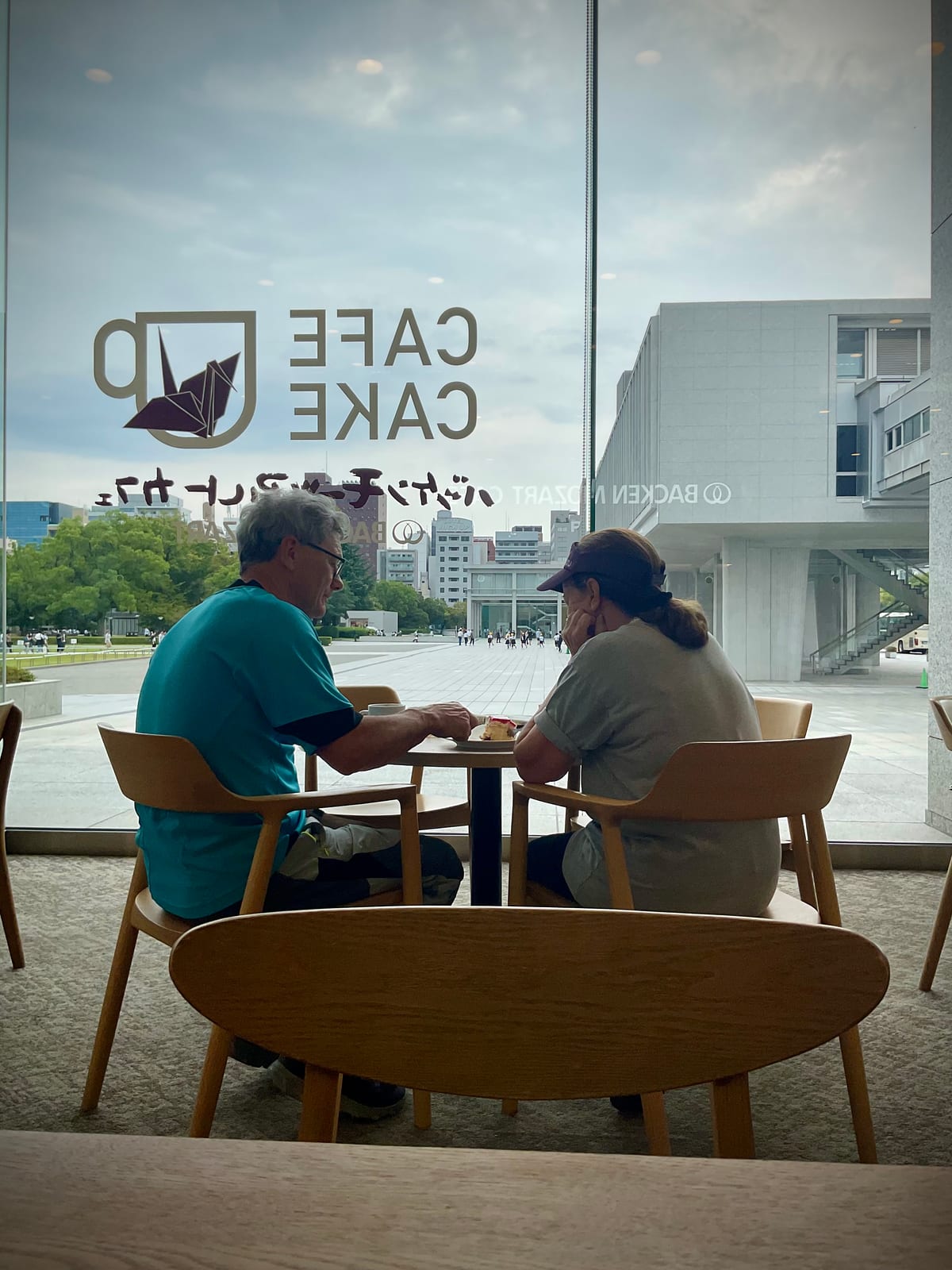 Seen through a cafe window, two people sit at a table, one turned away, the glass catching reflections of the street.