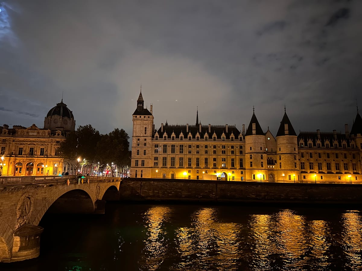 A turreted riverside palace lit at night beside an arched bridge, reflections breaking on the river.