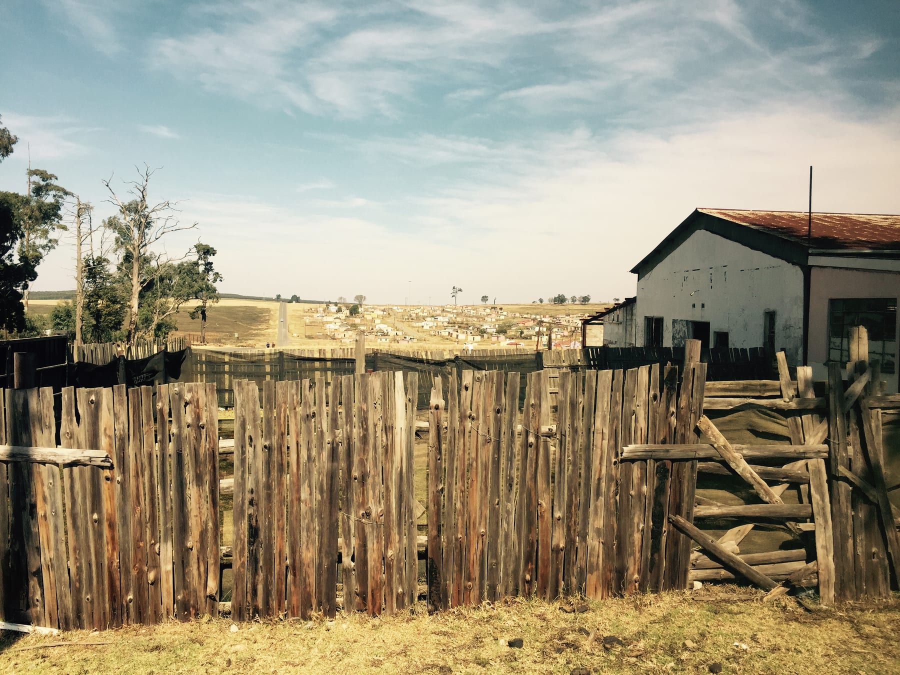 A rough timber stockade fence beside a simple building under a wide sky, red earth underfoot.