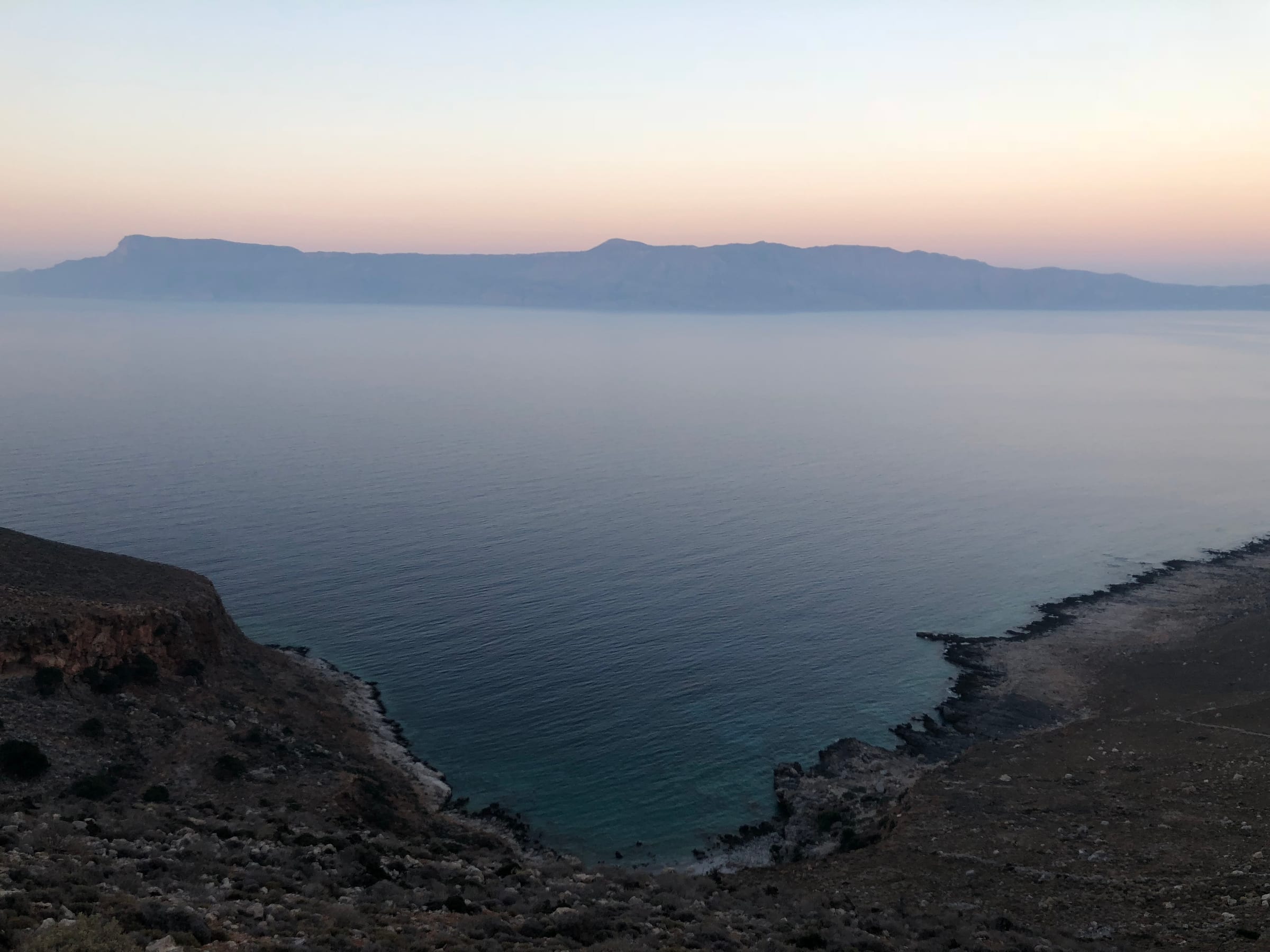 A hazy coastal headland and calm sea under soft pastel light, the horizon almost dissolved in mist.