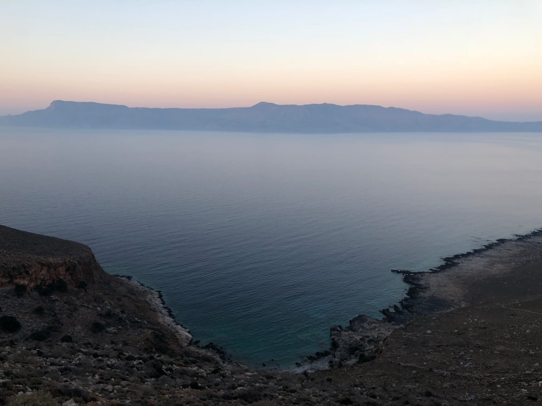 A hazy coastal headland and calm sea under soft pastel light, the horizon almost dissolved in mist.
